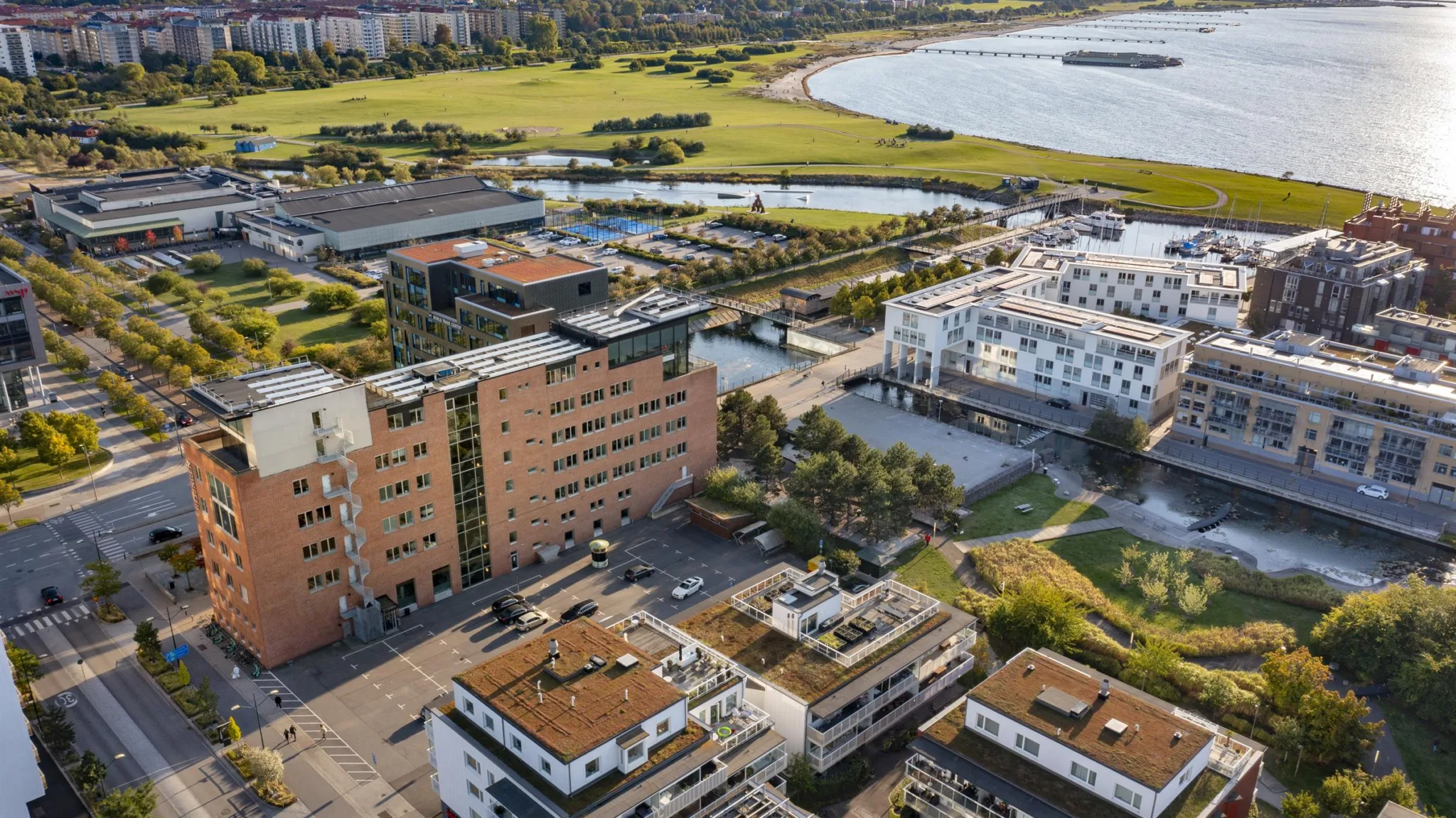 4. Flygbild över ett bostadsområde med byggnader, grönområden och en sjö i bakgrunden.