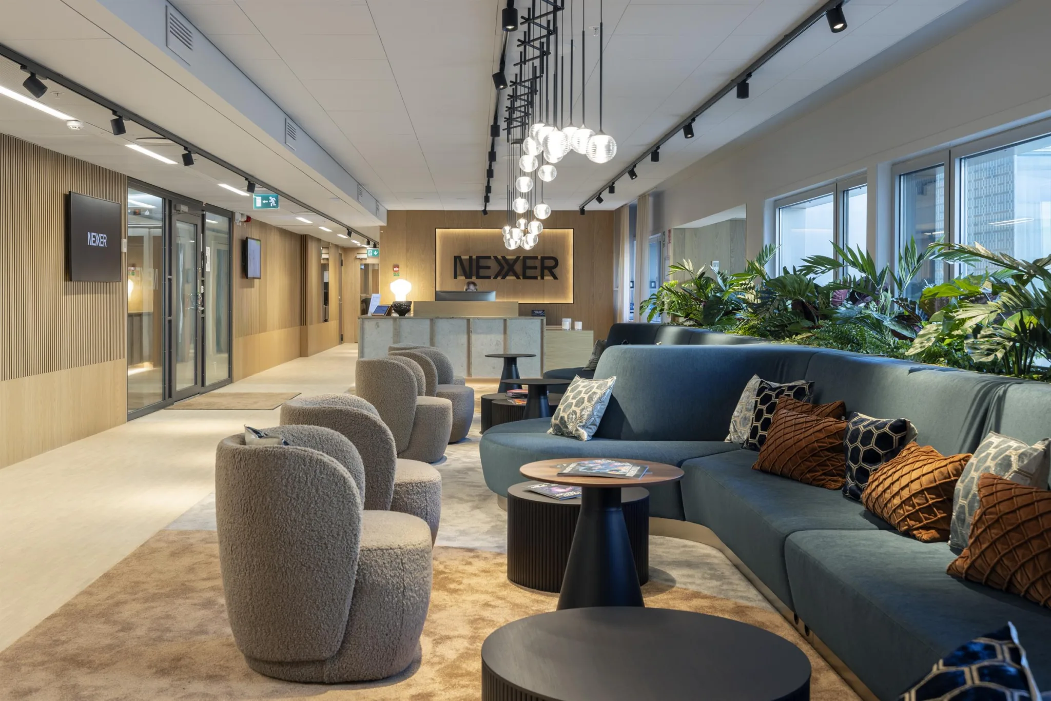 2. Ett stilrent loungeområde med soffor, fåtöljer, runda bord och växter, samt belysning i taket och en reception i bakgrunden.