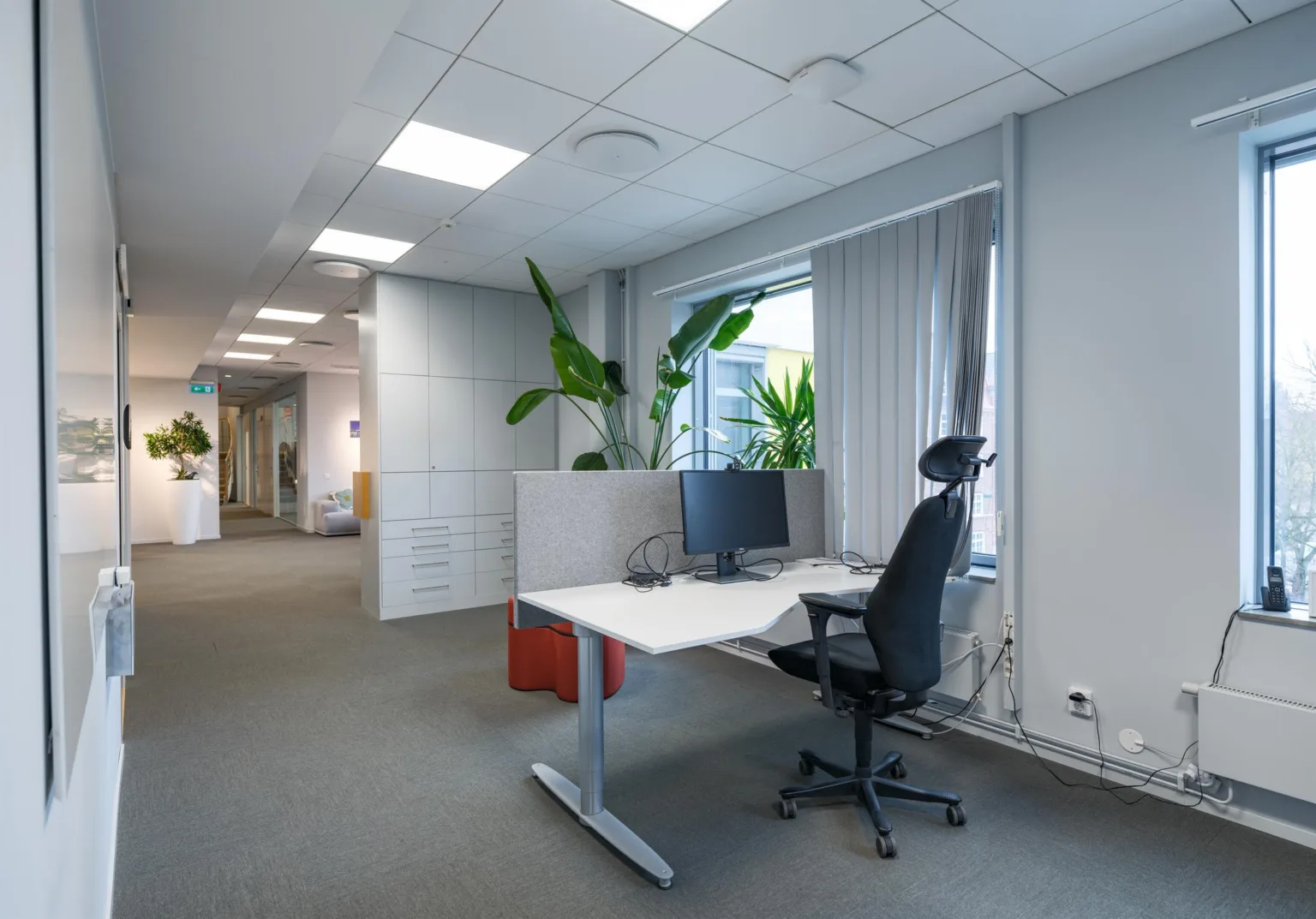 Arbetsstation med skrivbord, kontorsstol, datorskärm och växter i närheten av ett fönster.