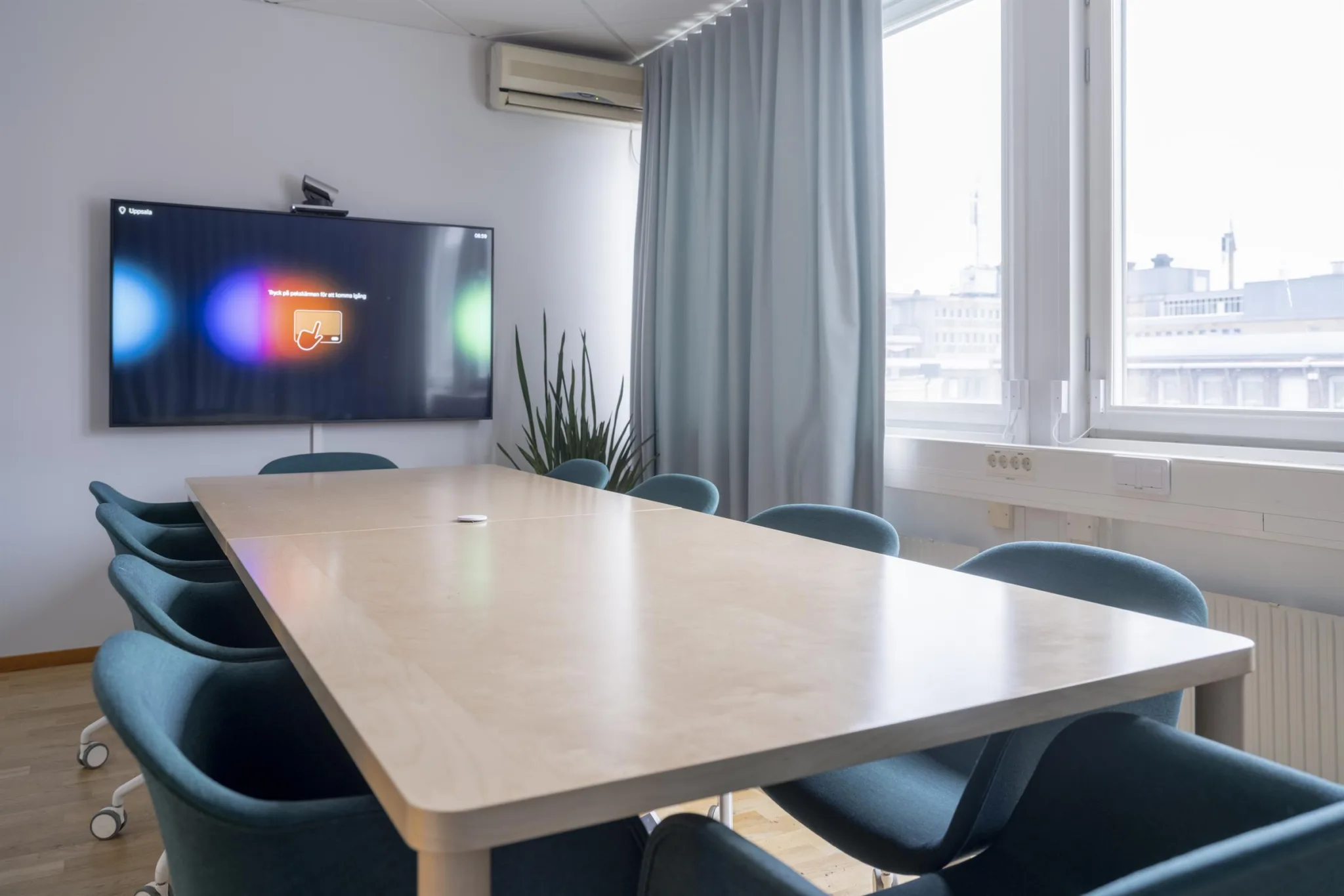 Ett mötesrum med ett långt bord, blå stolar, en TV på väggen, grå gardiner och ett stort fönster.