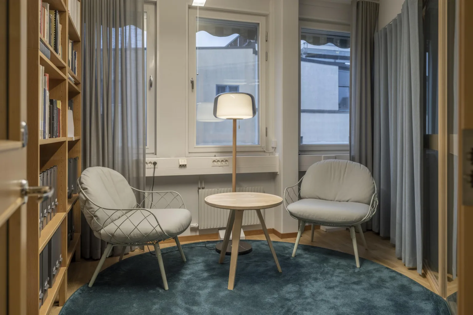 Två moderna fåtöljer med ett litet träbord och en golvlampa mellan dem, placerade på en blå matta vid fönster.