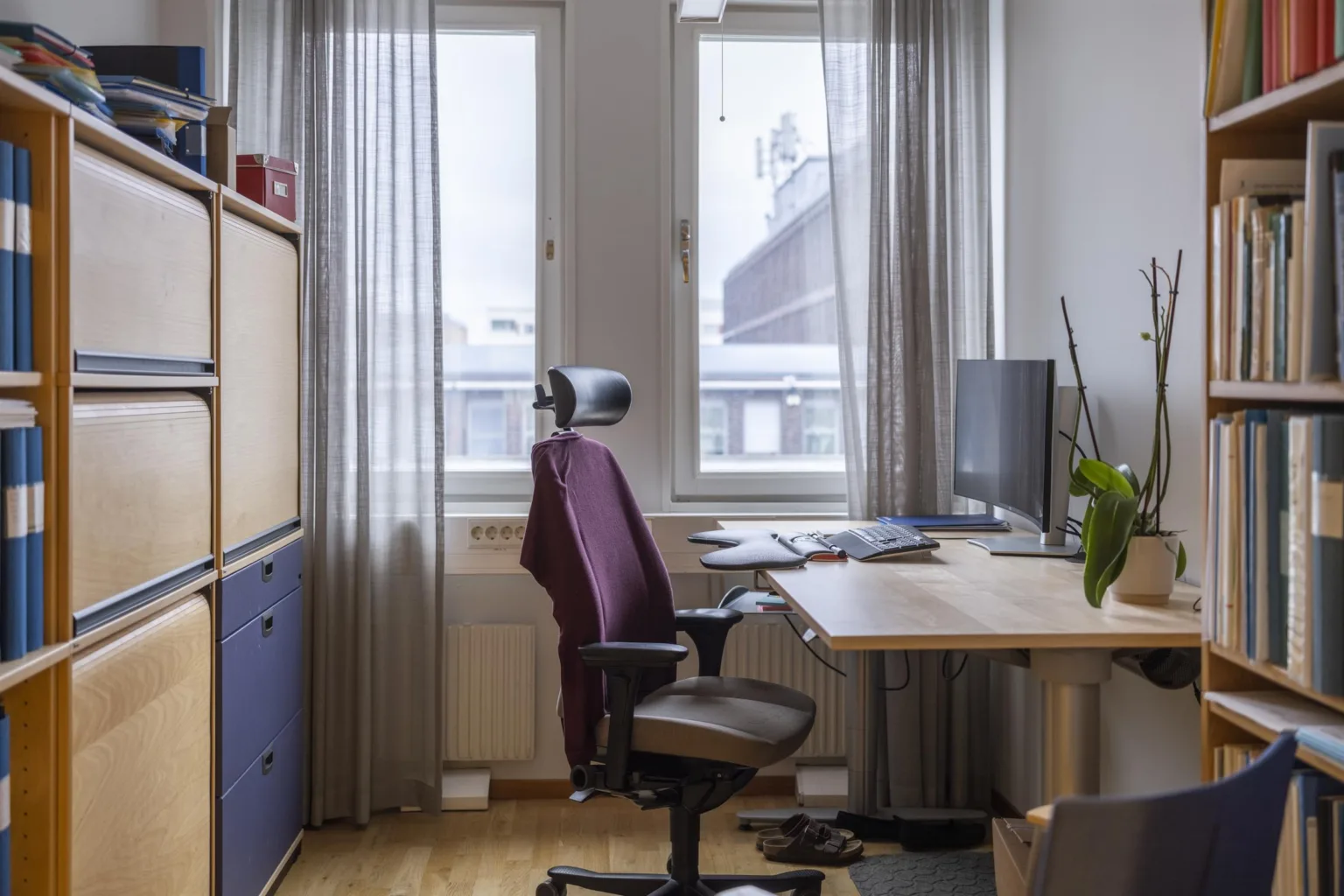 Ett hemmakontor med skrivbord, dator, ergonomisk stol, hylla och fönster med ljusa gardiner.