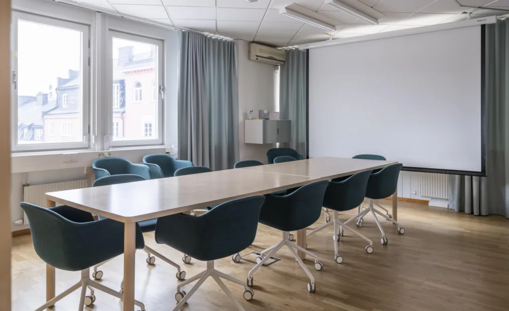 Ett konferensrum med ett långt bord, blå stolar på hjul, grå gardiner och ett stort fönster.