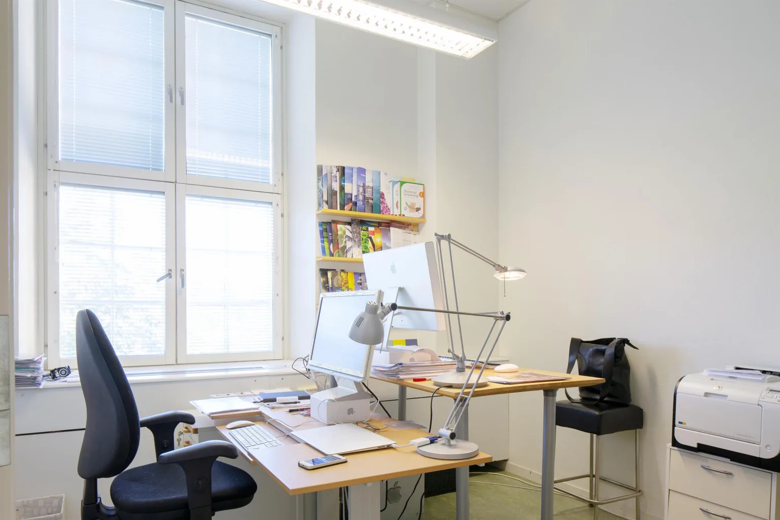 Ett kontor med ett skrivbord, en dator, skrivbordslampor och en hylla med böcker vid ett stort fönster.