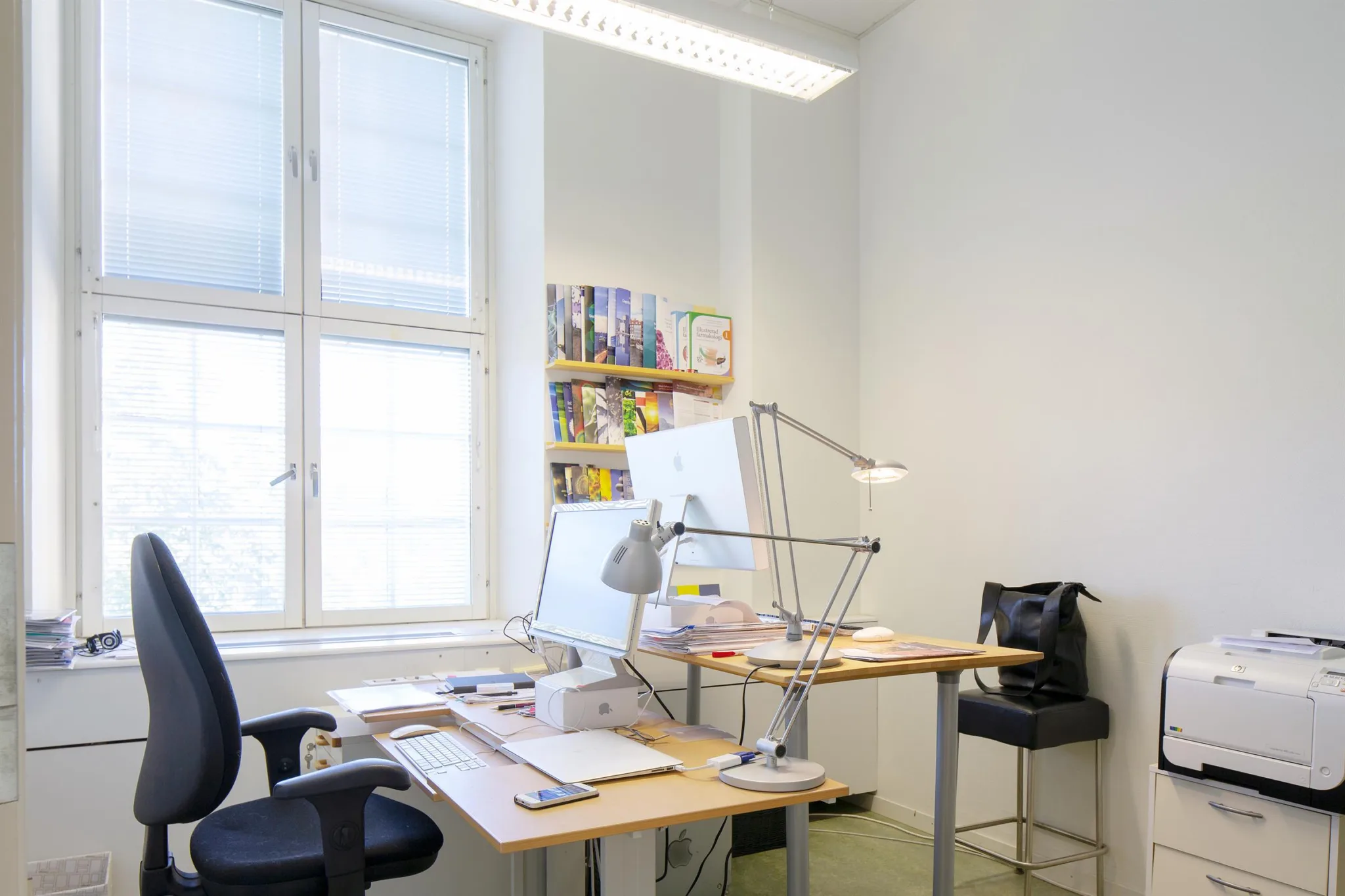 Ett kontor med ett skrivbord, en dator, skrivbordslampor och en hylla med böcker vid ett stort fönster.