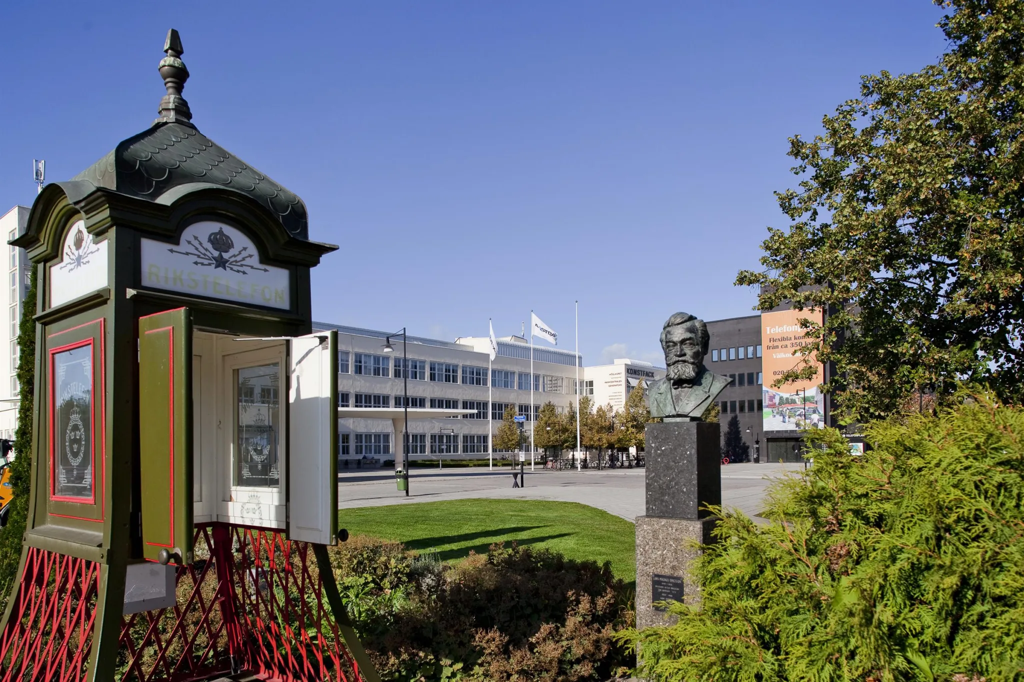 En utomhusmiljö med en byst på en piedestal, gröna buskar och ett öppet skåp bredvid, med en modern byggnad i bakgrunden.