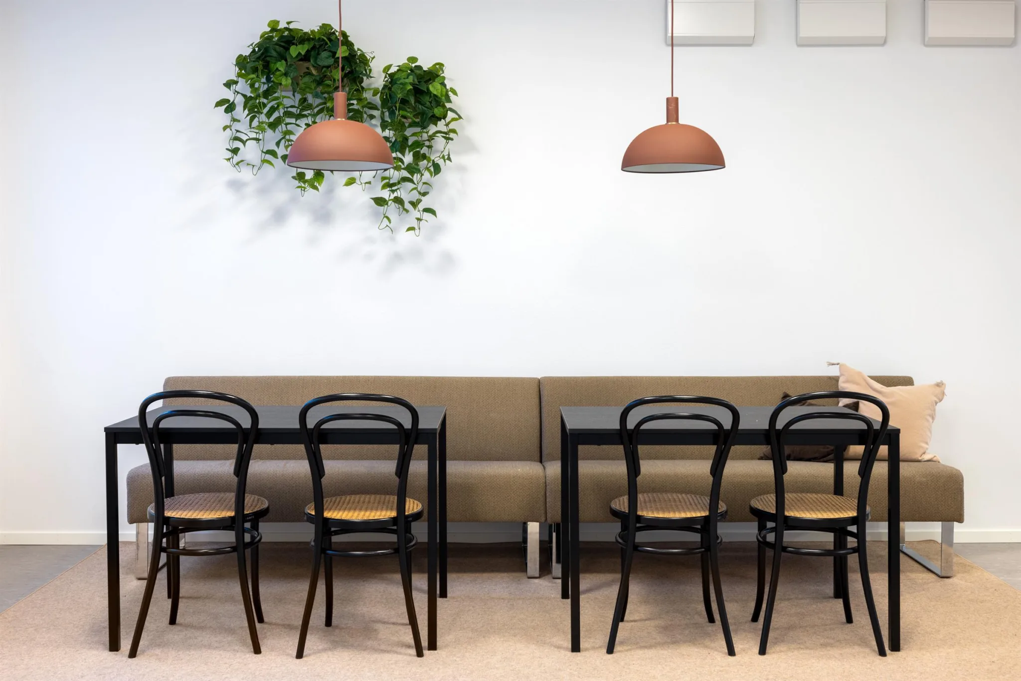 Ett matsalsområde med två svarta bord, stolar, beige bänk, gröna växter på väggen och två taklampor.