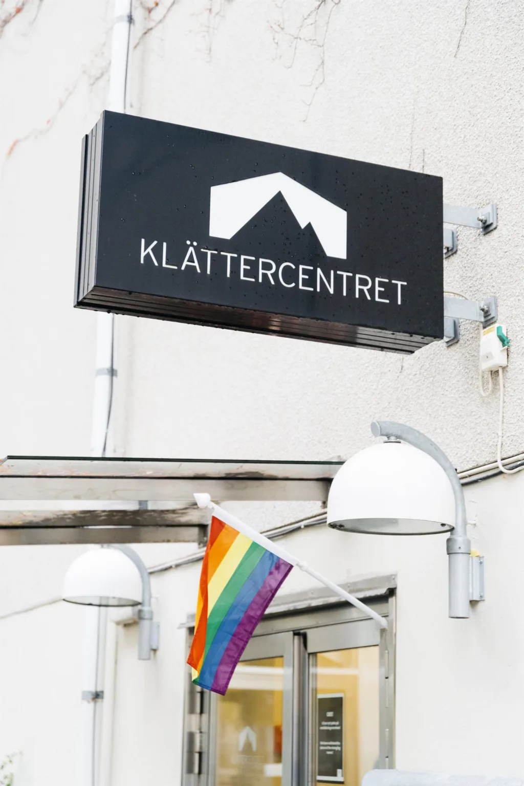 En svart skylt med texten "Klättercentret" och en regnbågsflagga hängande utanför en byggnad.