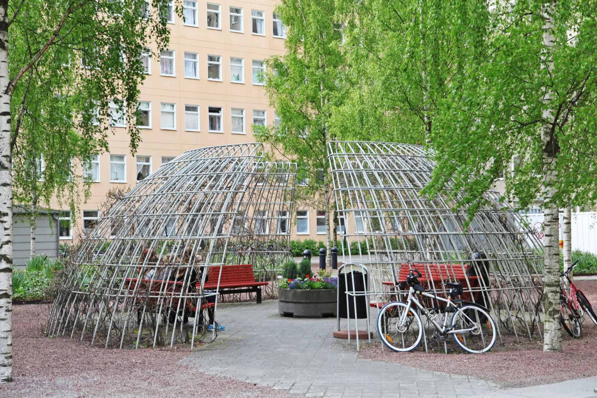 En utomhusplats med ett gallerformigt tak, röda bänkar, en cykel och träd i bakgrunden.
