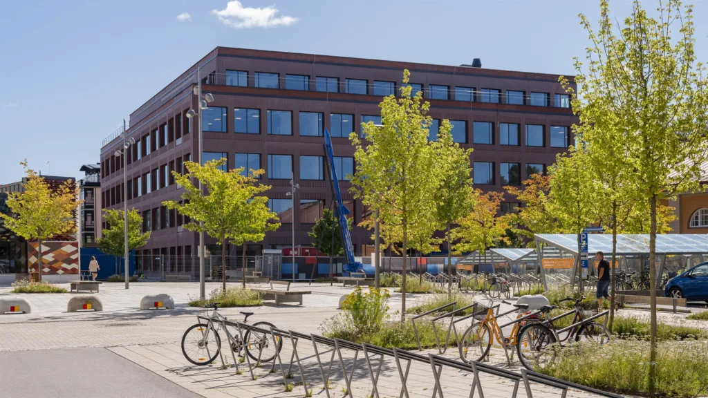 En modern byggnad i rött tegel med fönster, omgiven av cykelställ, träd och en öppen plats.