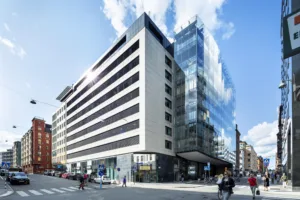 En modern byggnad med glasfasad och vita paneler, omgiven av en stadsmiljö med gående och bilar.