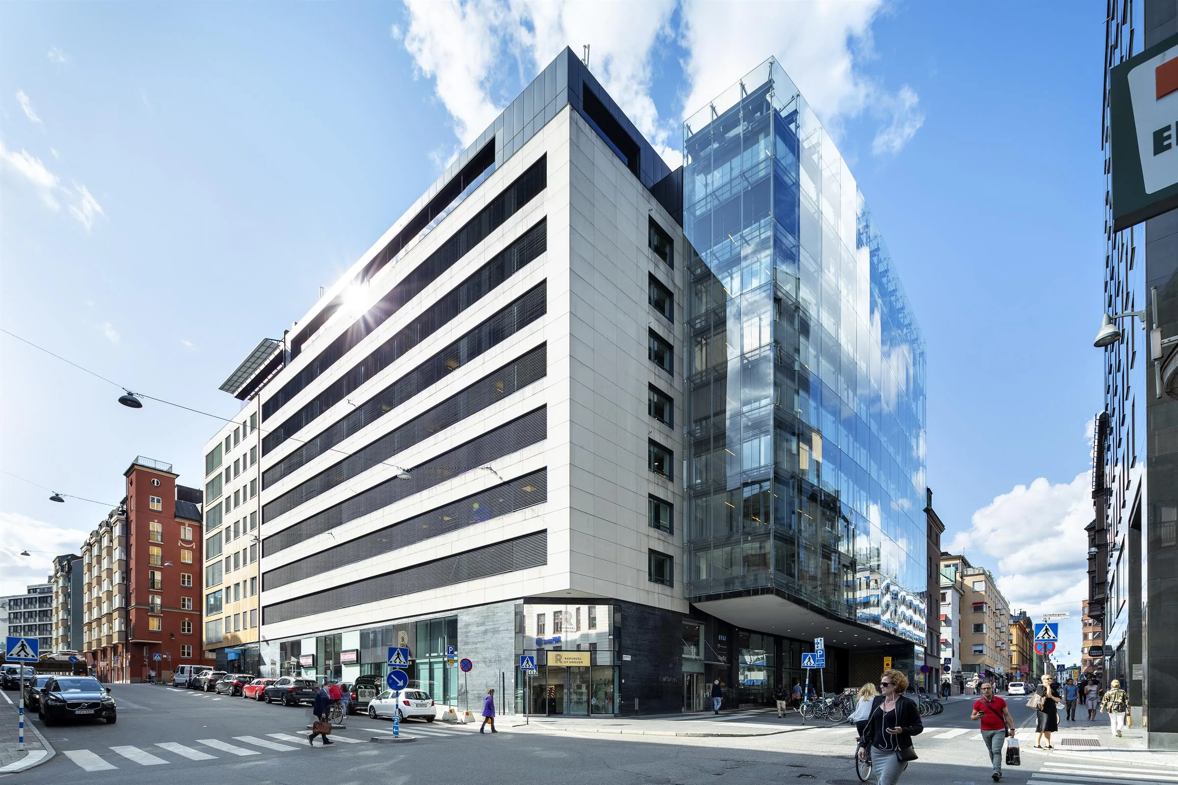 En modern byggnad med glasfasad och vita paneler, omgiven av en stadsmiljö med gående och bilar.