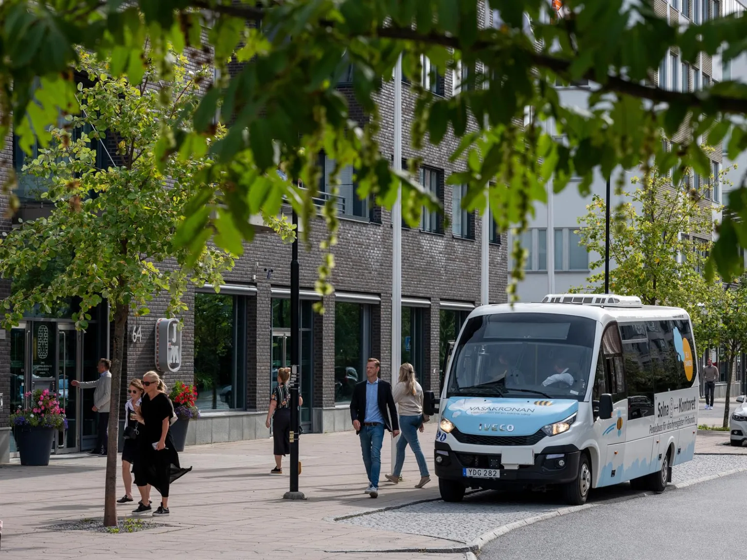 En liten blå och vit buss kör förbi en gata med träd och fotgängare.