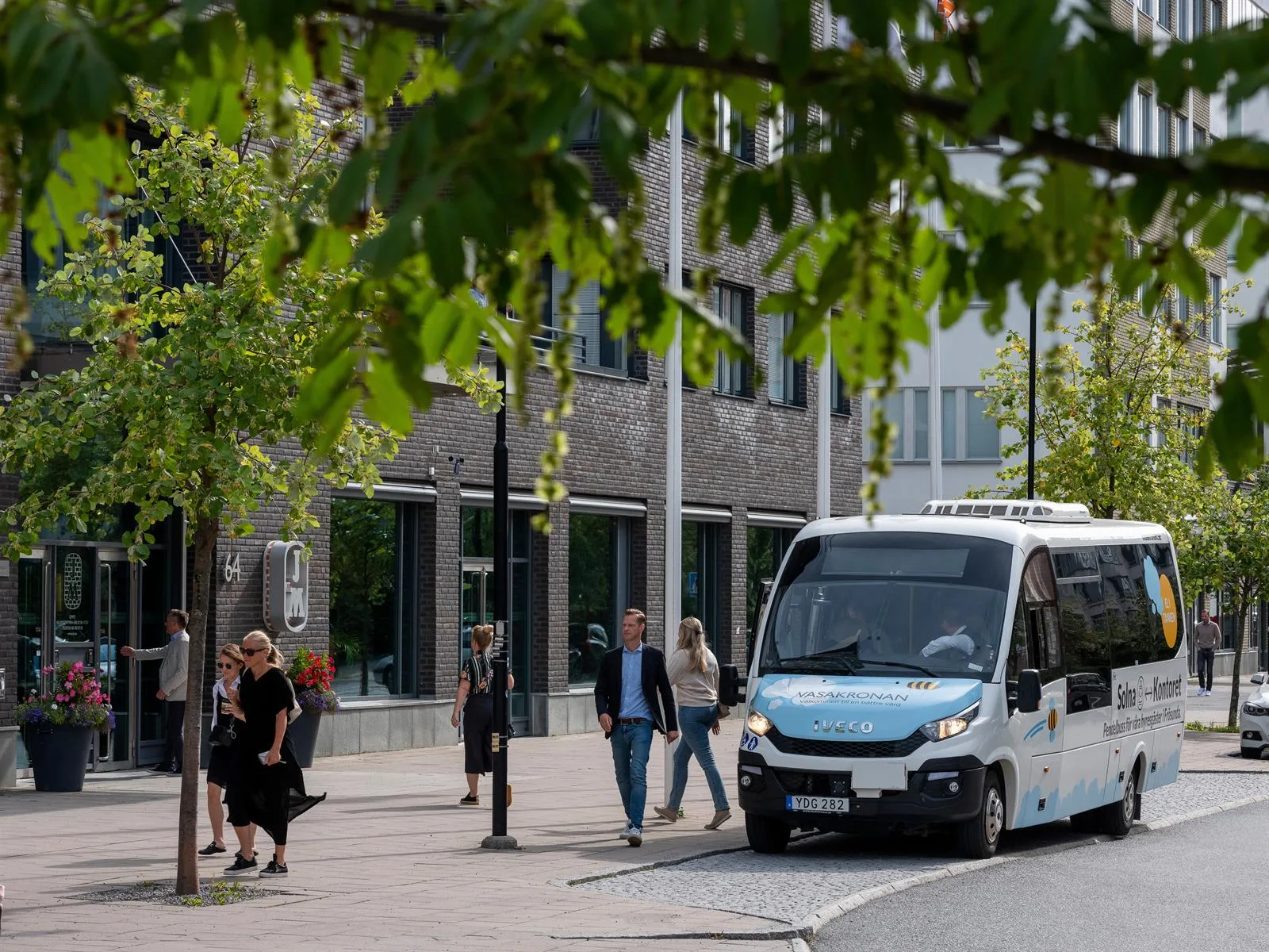 En liten blå och vit buss kör förbi en gata med träd och fotgängare.