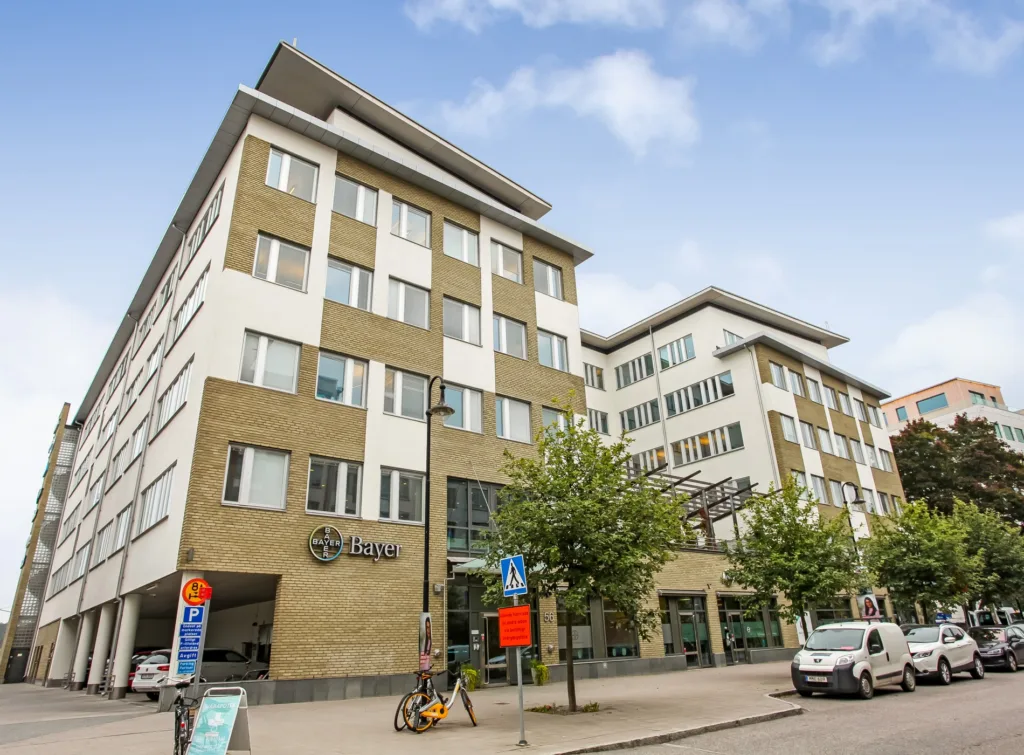 Ett modernt flerbostadshus med en kombination av vita och beige fasader, omgivna av träd och parkerade bilar.