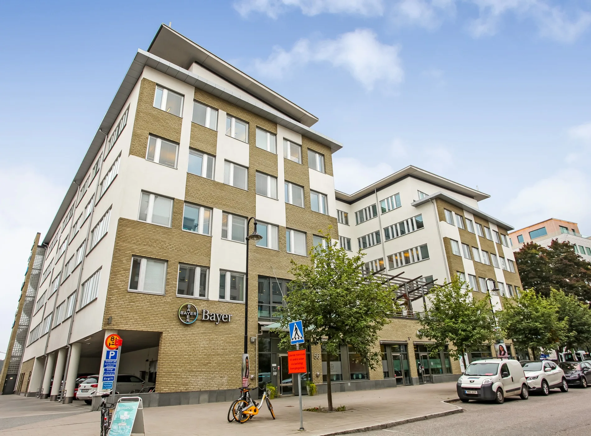 Ett modernt flerbostadshus med en kombination av vita och beige fasader, omgivna av träd och parkerade bilar.
