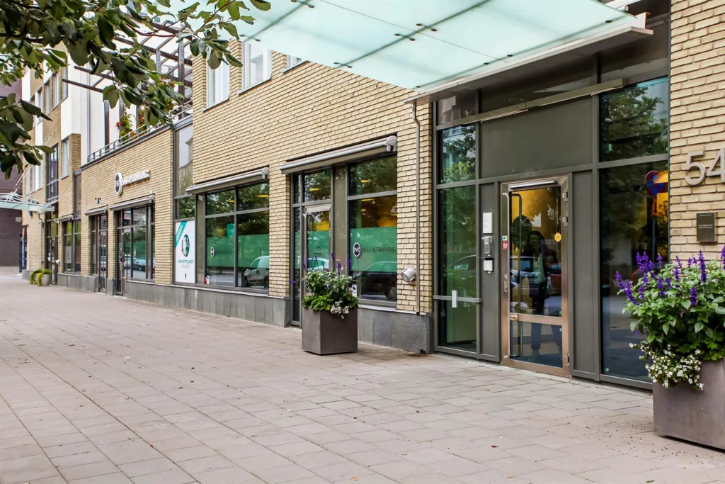 Entré till en byggnad med glasdörrar och stora fönster, omgiven av en gångväg med planteringar.