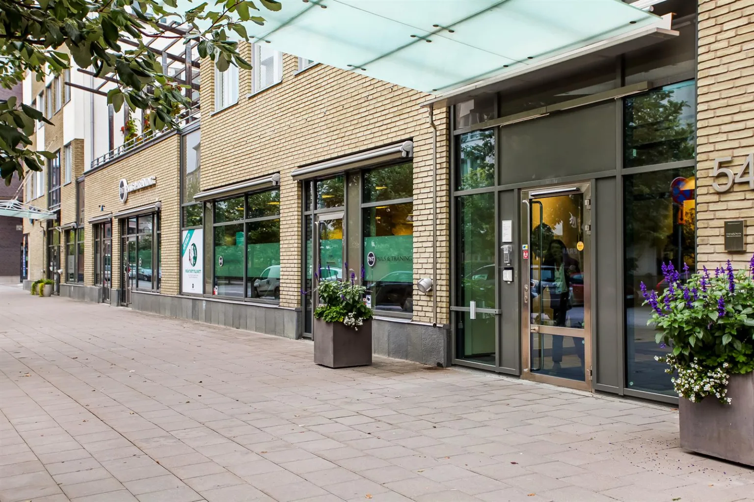 Entré till en byggnad med glasdörrar och stora fönster, omgiven av en gångväg med planteringar.
