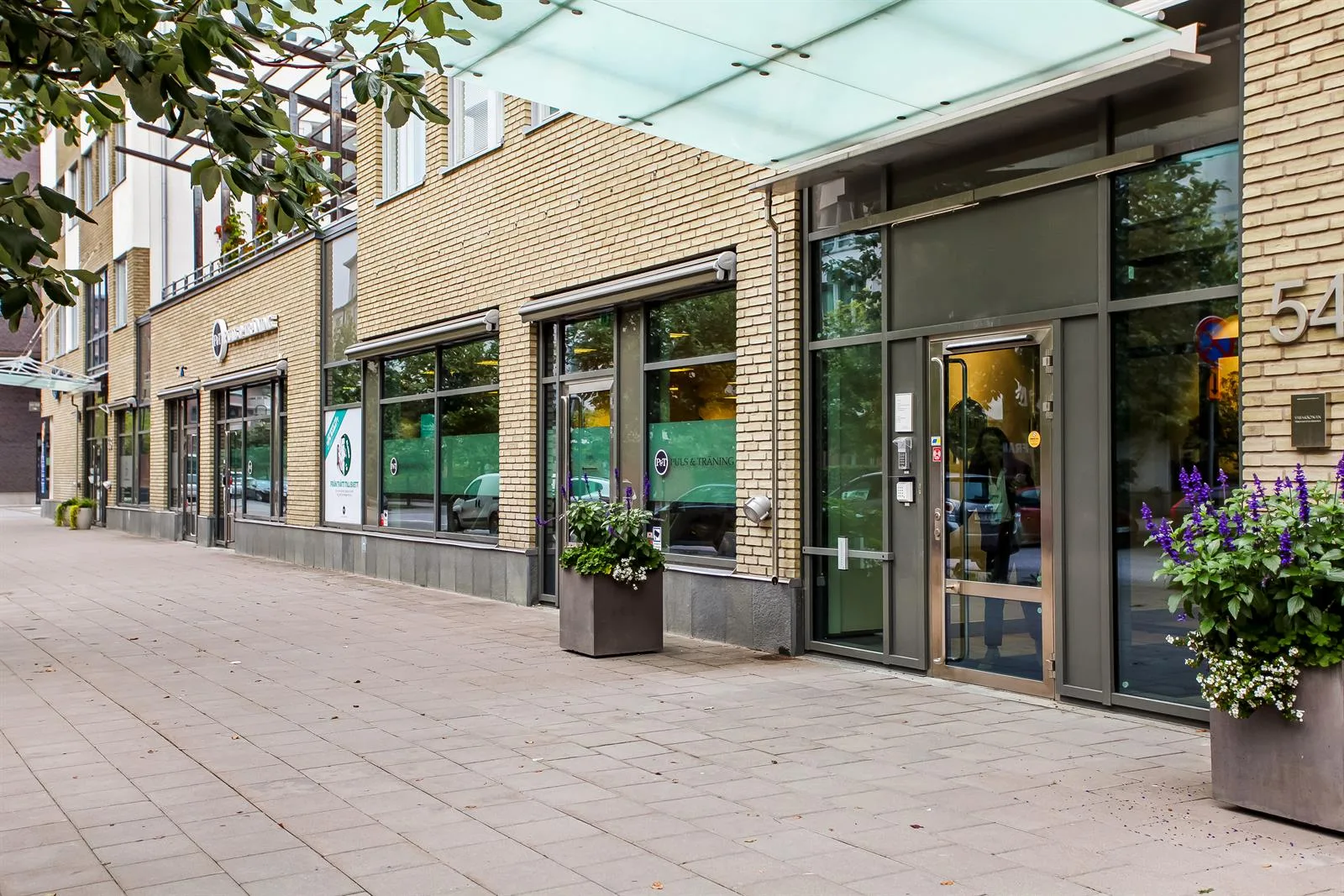 Entré till en byggnad med glasdörrar och stora fönster, omgiven av en gångväg med planteringar.