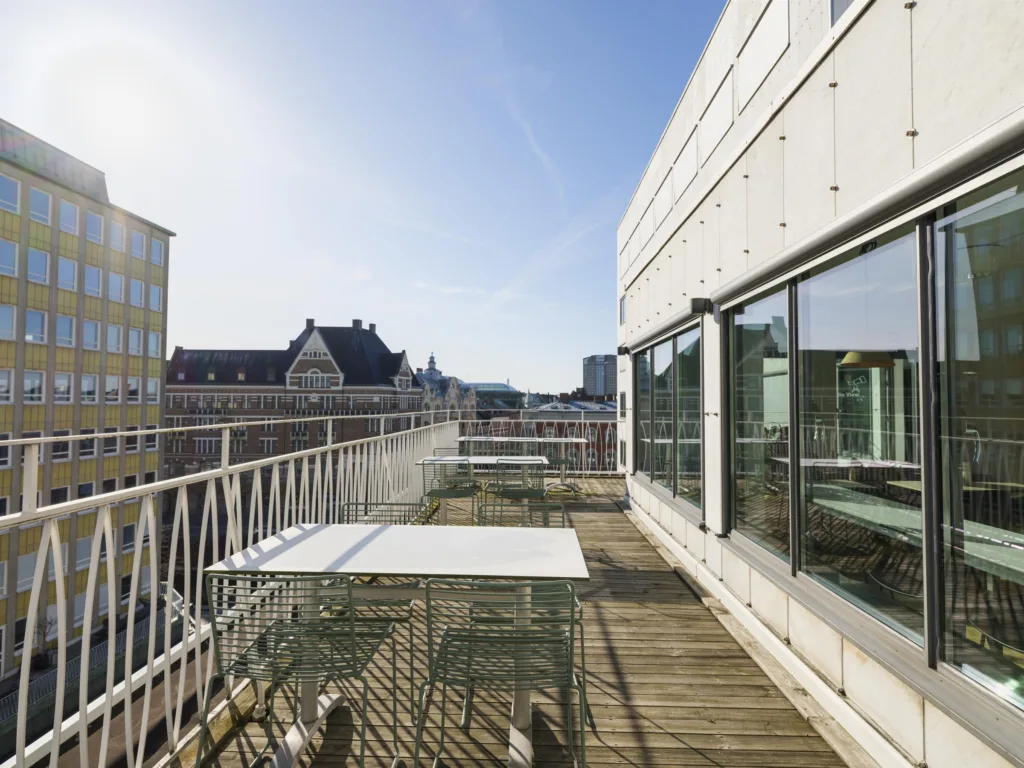En möblerad takterrass med vita bord och stolar, omgiven av glasfasader och en vy över stadens byggnader.