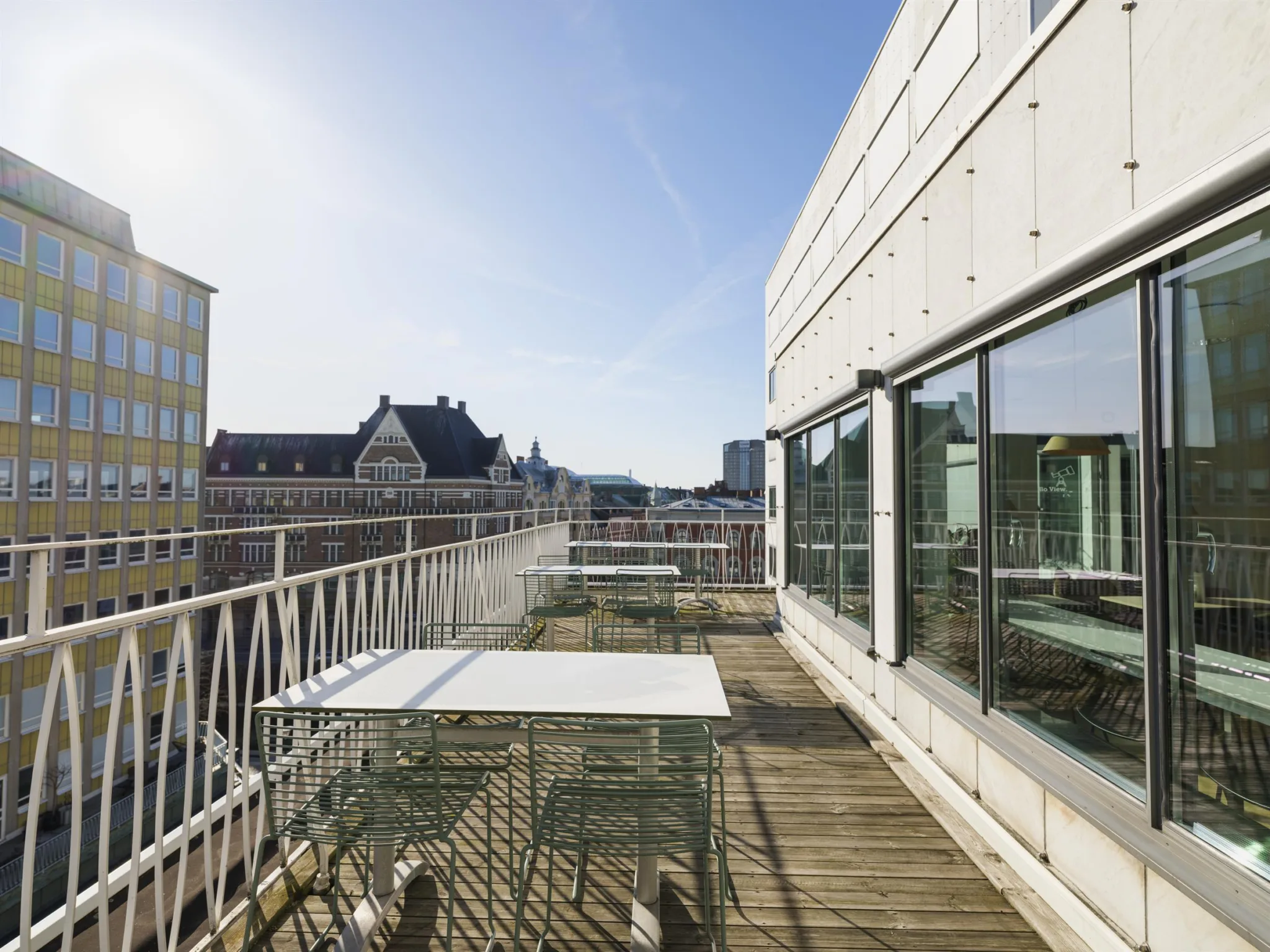 En möblerad takterrass med vita bord och stolar, omgiven av glasfasader och en vy över stadens byggnader.