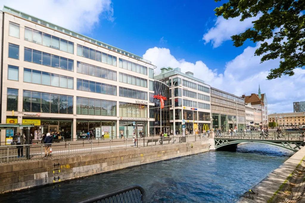 En modern byggnad med glasfasad vid en kanal, med bro och vatten under en solig himmel.