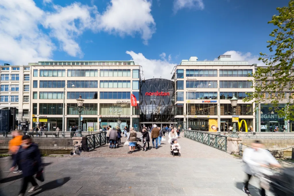 2. Entré till köpcentrum med glasfasad, omgiven av människor och flaggor under en klarblå himmel.