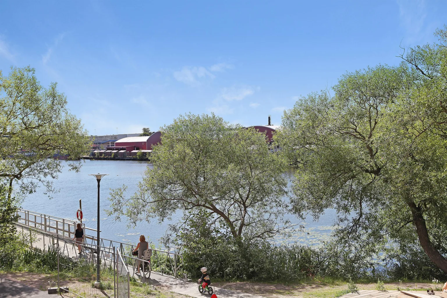 En sjö med träd i förgrunden och röda byggnader på andra sidan vattnet under en klar blå himmel.