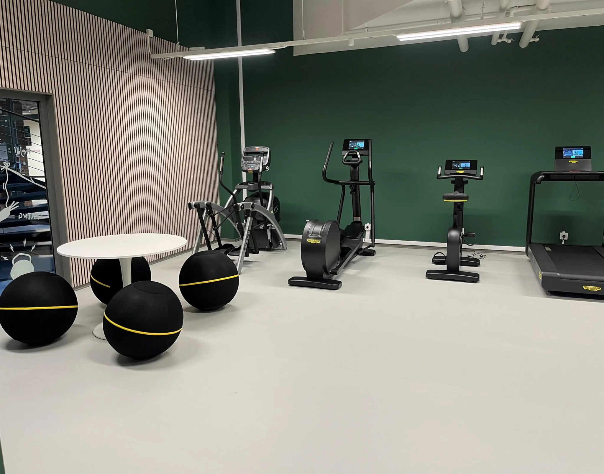 Ett gymrum med gröna väggar, träningsutrustning inklusive motionscyklar, roddmaskin och bollar samt ett bord.