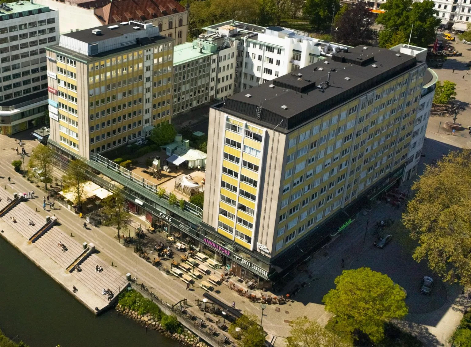 En flygbild av en modern byggnad med en grönskande omgivning och uteserveringar vid en kanal.