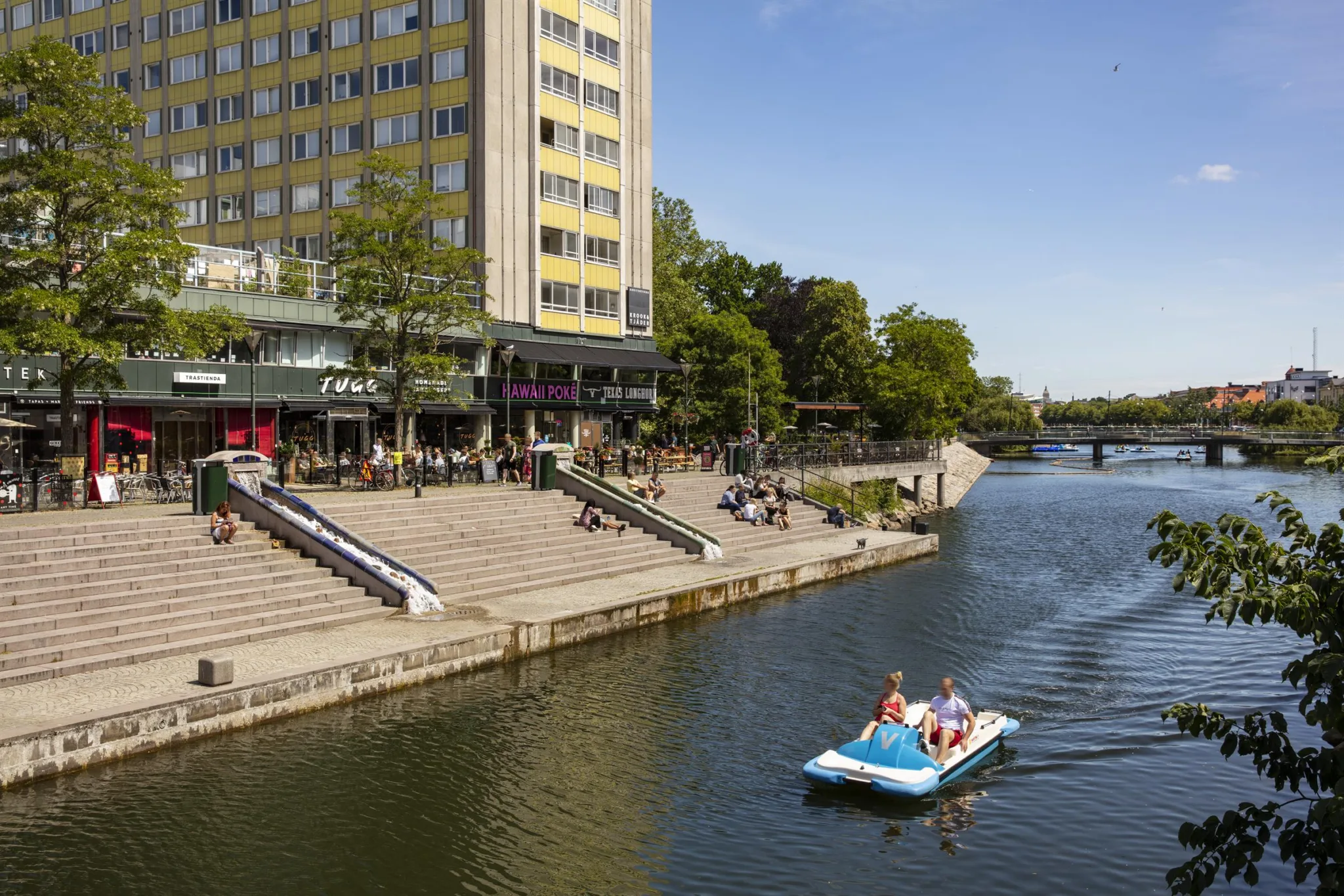 En kanal med en trampbåt i förgrunden, människor på trappor vid vattnet och en stor byggnad i bakgrunden.