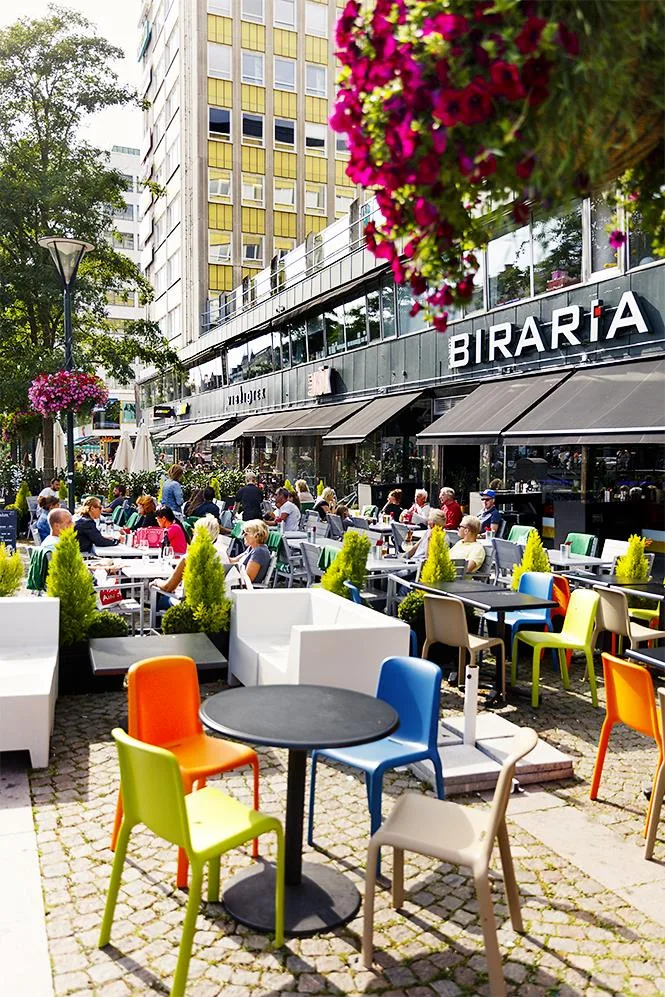 En uteservering med färgglada stolar och bord framför restaurangen Biraria, omgiven av blommor och grönska.