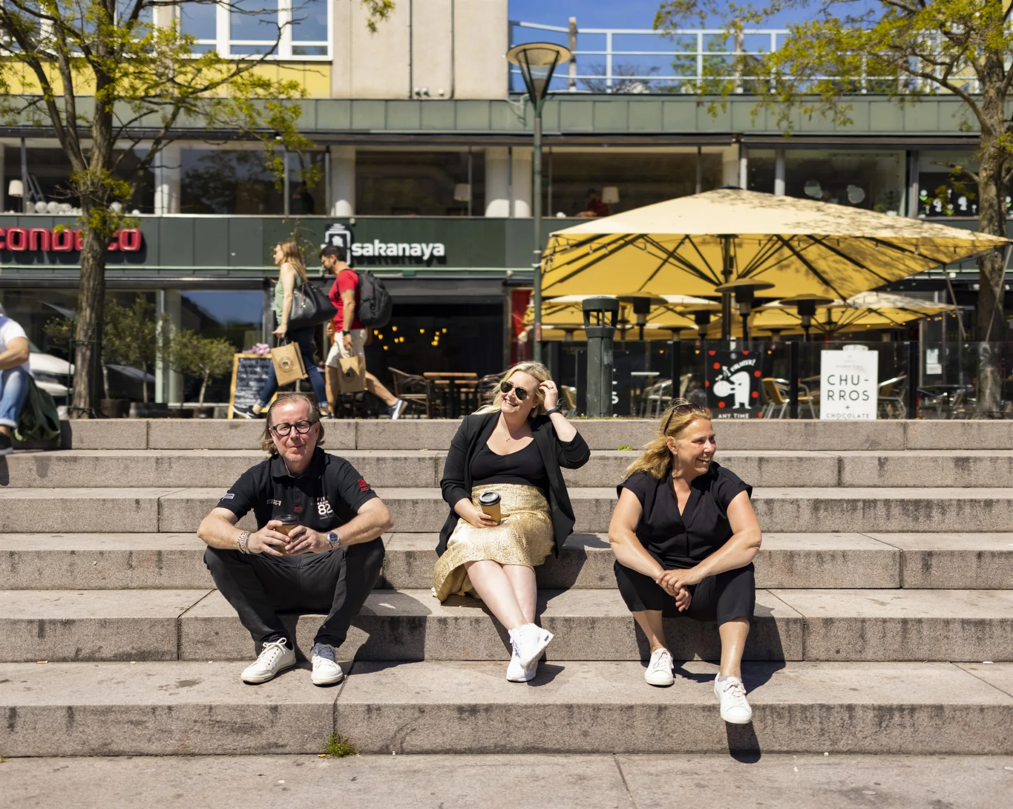 Tre personer sitter på trappor framför ett café med gula parasoller och människor i bakgrunden.