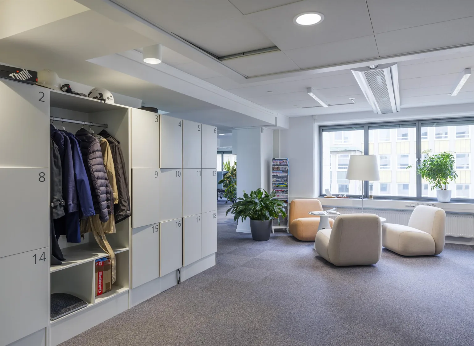 4. Ett kontorsutrymme med en klädförvaring, fåtöljer, en lampa och en stor grön växt bredvid ett fönster.