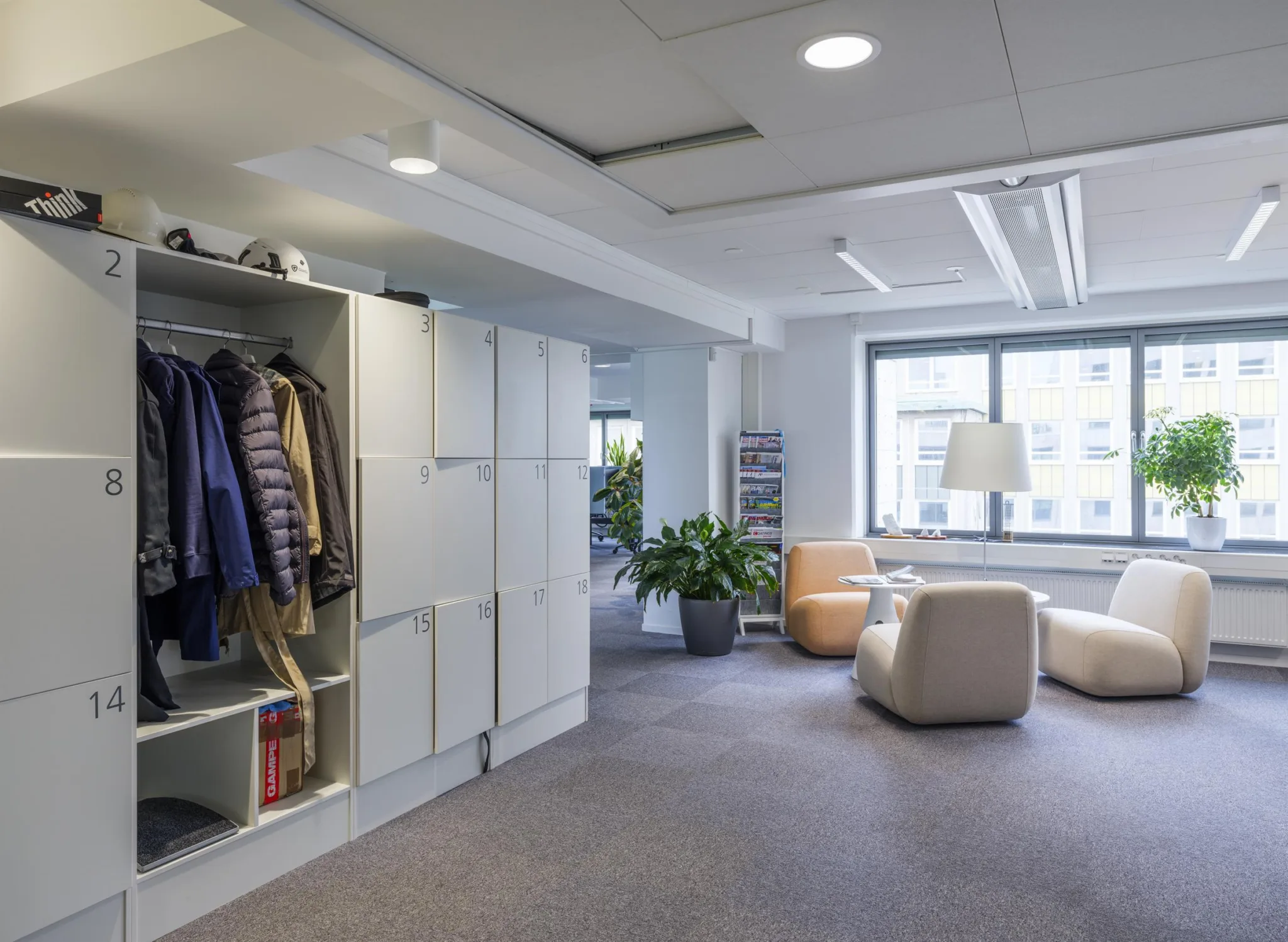 4. Ett kontorsutrymme med en klädförvaring, fåtöljer, en lampa och en stor grön växt bredvid ett fönster.