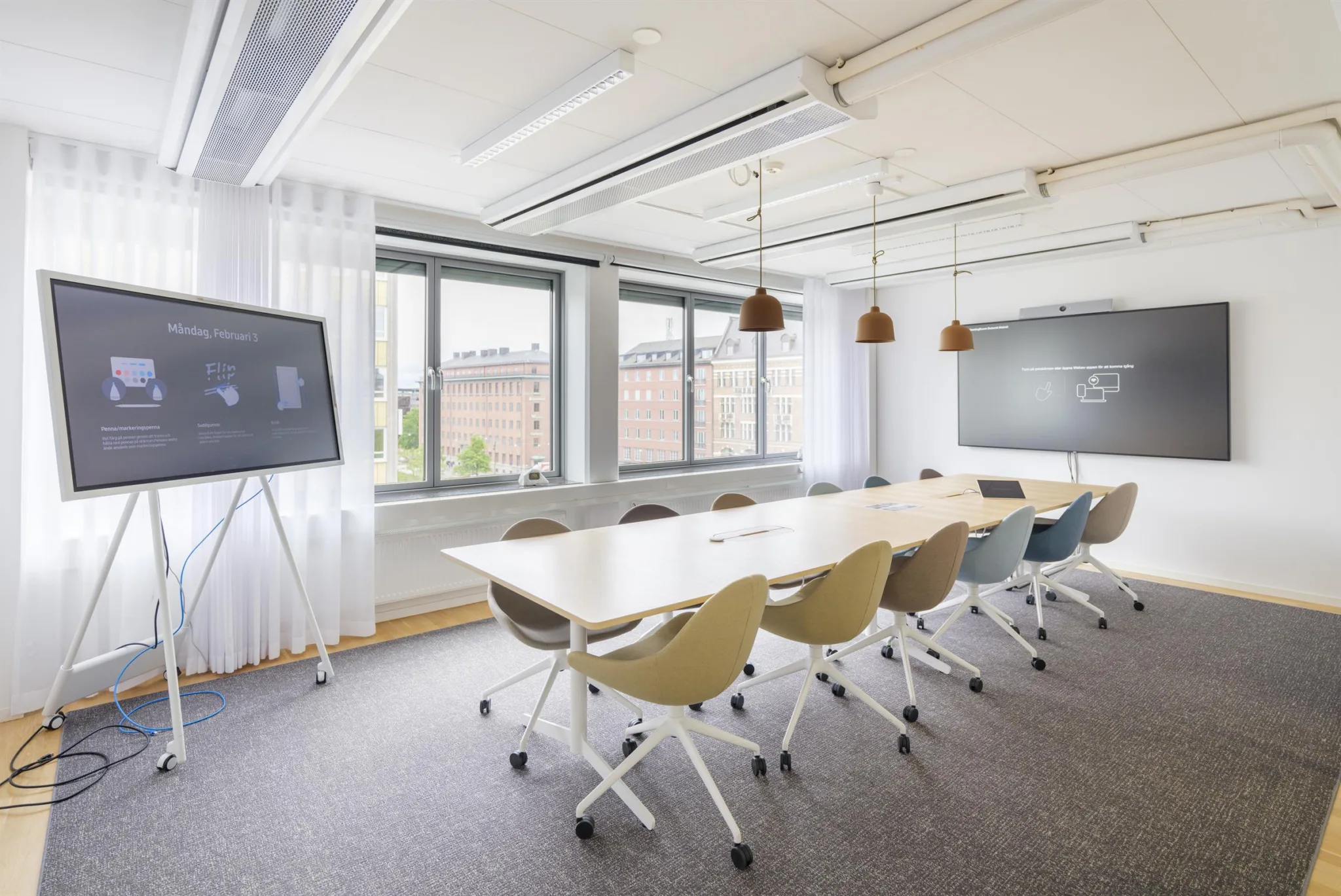 1. Ett modernt konferensrum med en lång rektangulär träbord, färgglada stolar på hjul och två stora fönster som släpper in naturligt ljus.