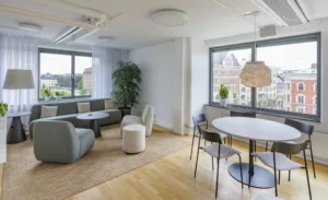 5. Ett ljust sällskapsrum med soffor, stolar, ett runt bord och stora fönster som vetter mot stadsvyer.