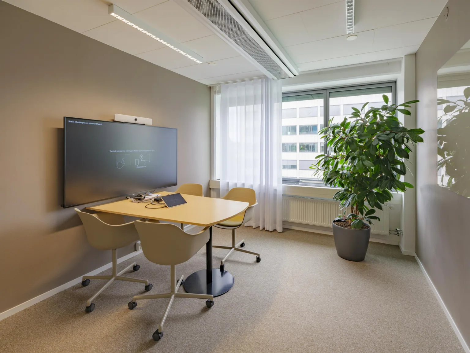 Ett mötesrum med ett litet bord, fyra stolar på hjul, en stor skärm på väggen och en grön växt bredvid ett fönster.