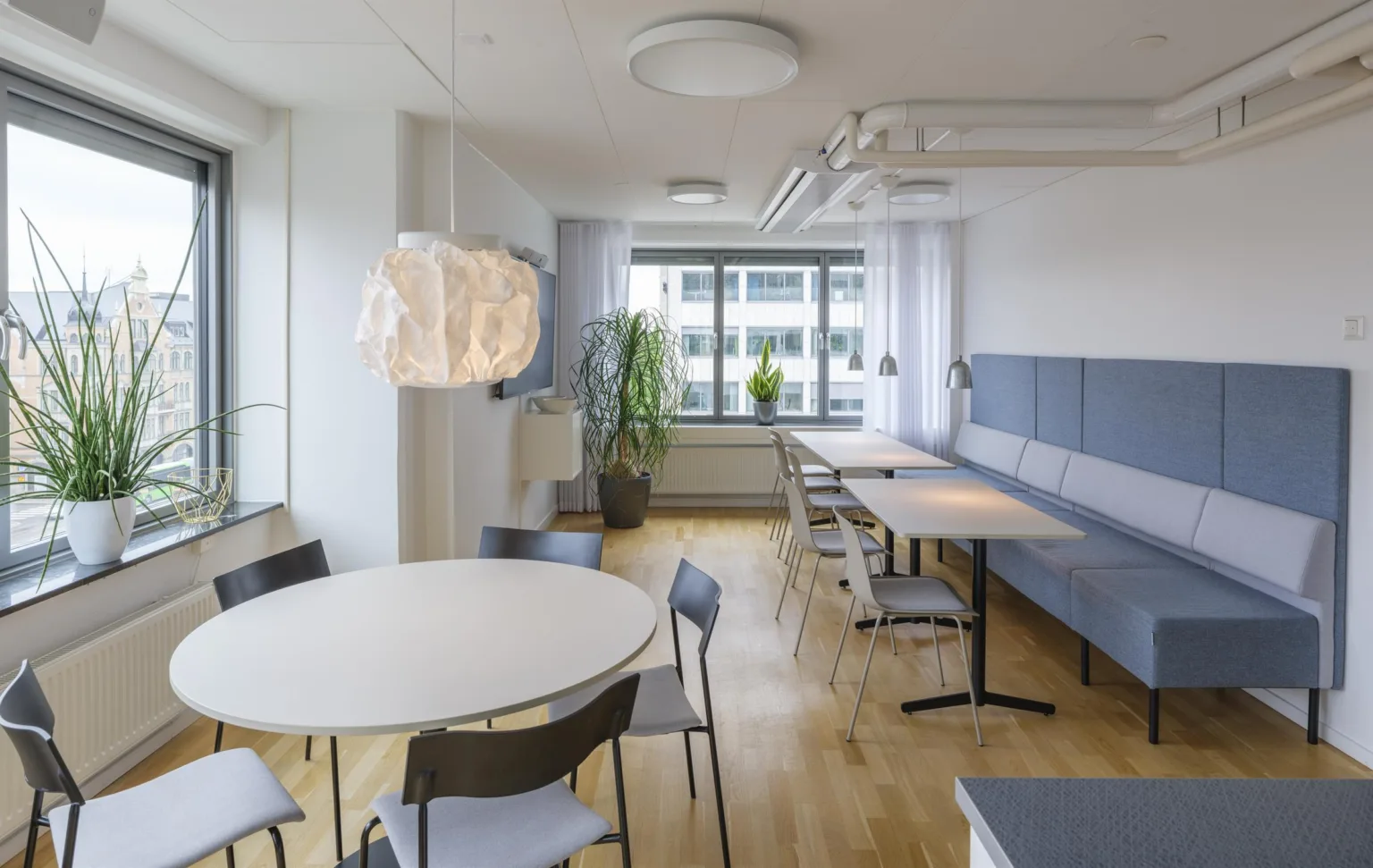 2. En ljus matsal med runda och rektangulära bord, stolar, en hängande lampa och växter vid fönstren.