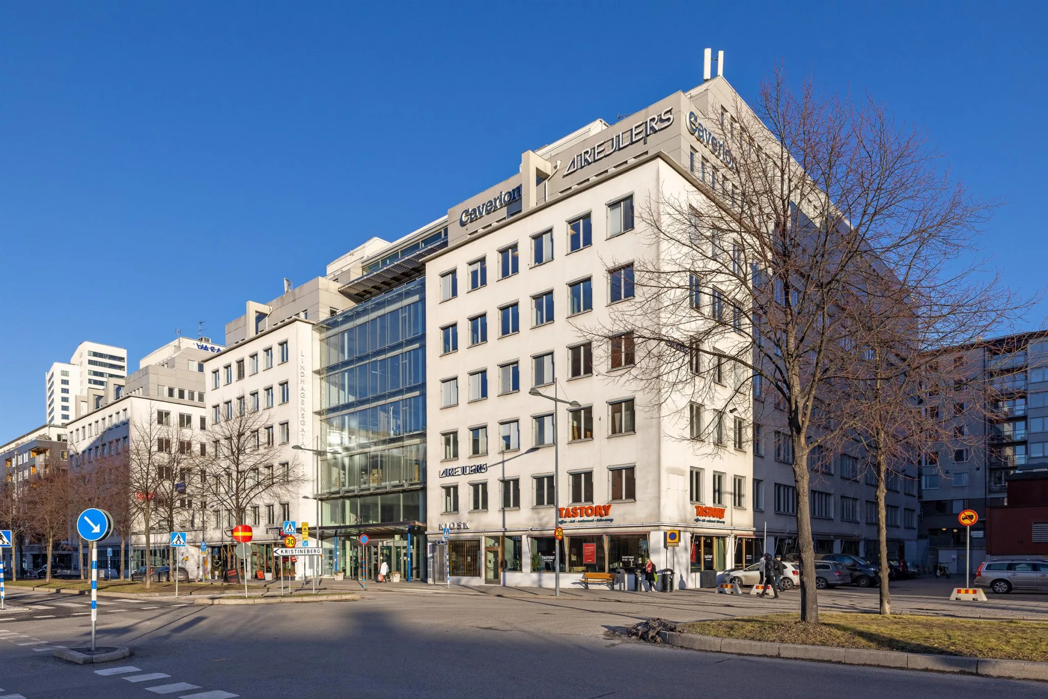 En modern byggnad med flera våningar, stora glaspartier och affärer på bottenvåningen. Träd och en trottoar syns i förgrunden.