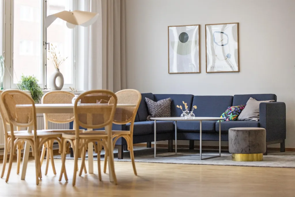 Ett vardagsrum med en matgrupp med rottingstolar, en mörkblå hörnsoffa, två tavlor på väggen och en modern inredning.