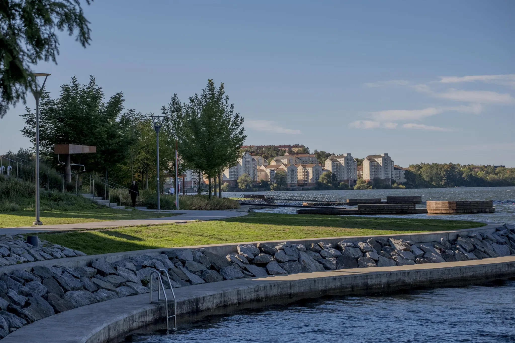 En park med stenmur, gröna träd och gräsmattor nära en vattendrag med stadshus i bakgrunden.