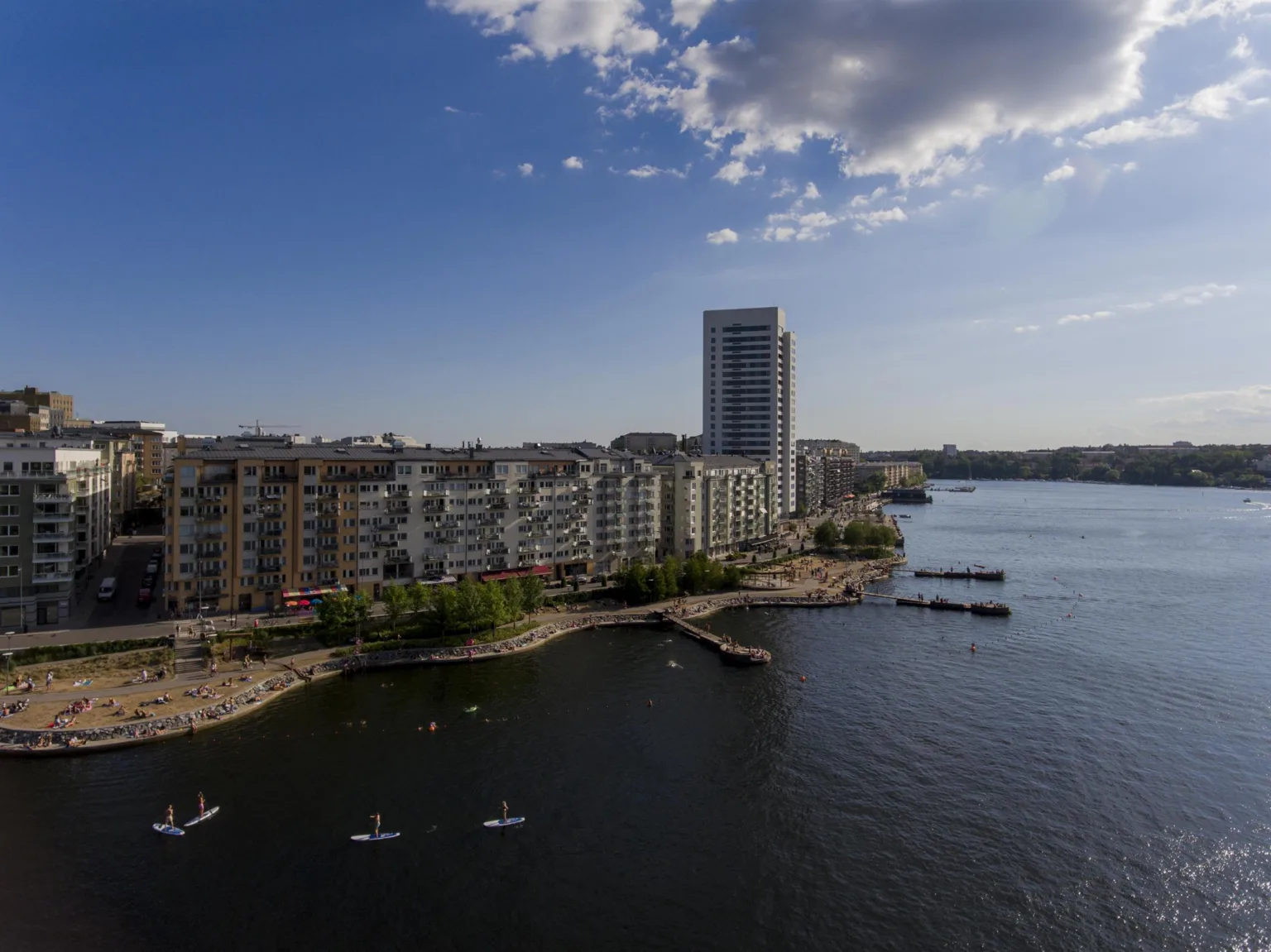 En stadsdel vid vatten med bostadshus, bryggor och små båtar under en solig himmel med moln.