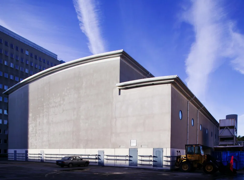 4. En stor byggnad med välvt tak mot en klar blå himmel och vita molnstrimmor, med bilar och en lastmaskin i förgrunden.