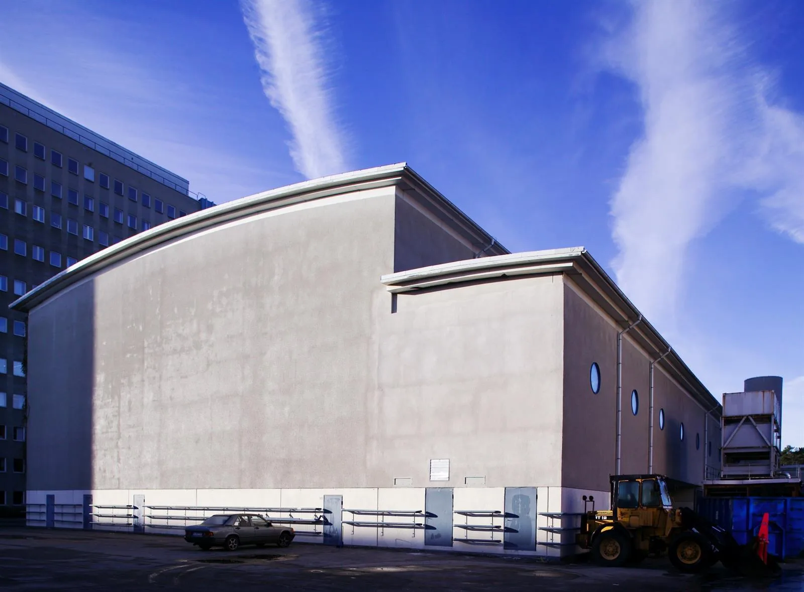 4. En stor byggnad med välvt tak mot en klar blå himmel och vita molnstrimmor, med bilar och en lastmaskin i förgrunden.