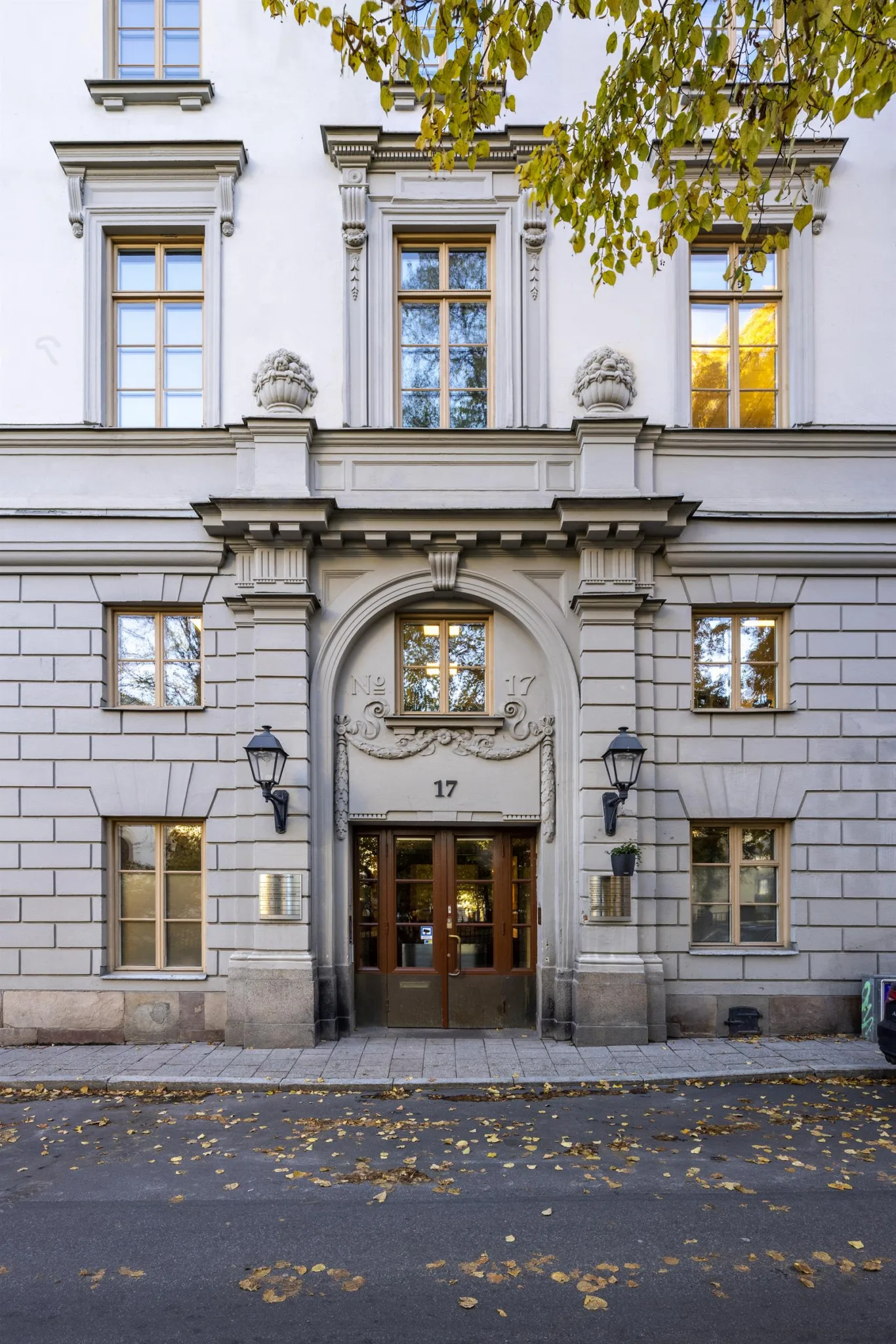 En byggnadsfasad med en stor, utsmyckad entré, två lyktstolpar och fönster med trädetaljer.