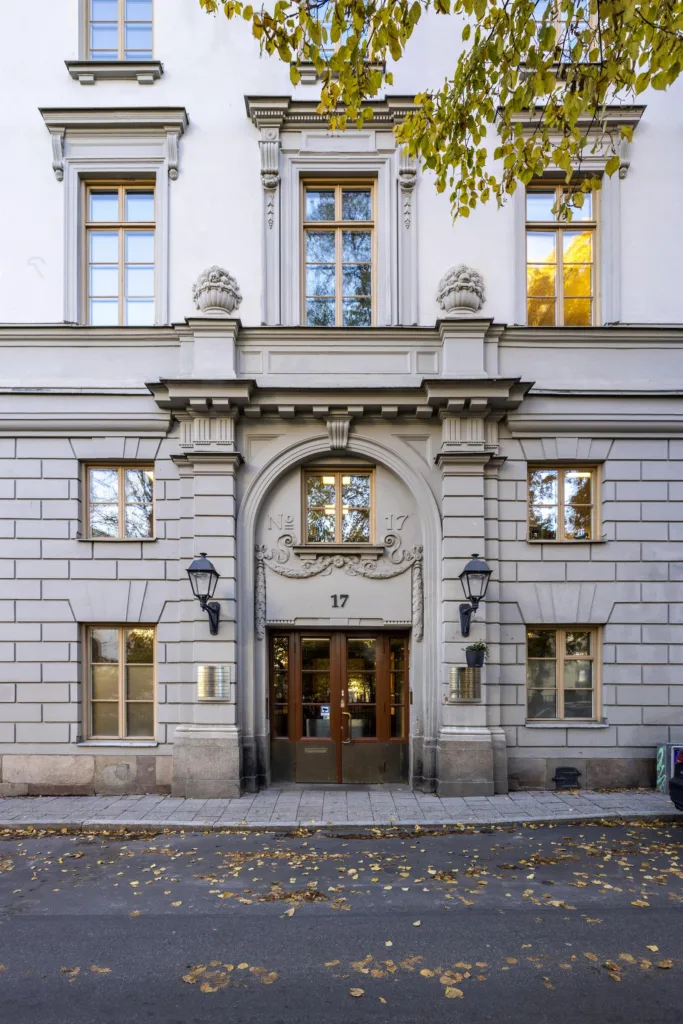 En byggnadsfasad med en stor, utsmyckad entré, två lyktstolpar och fönster med trädetaljer.