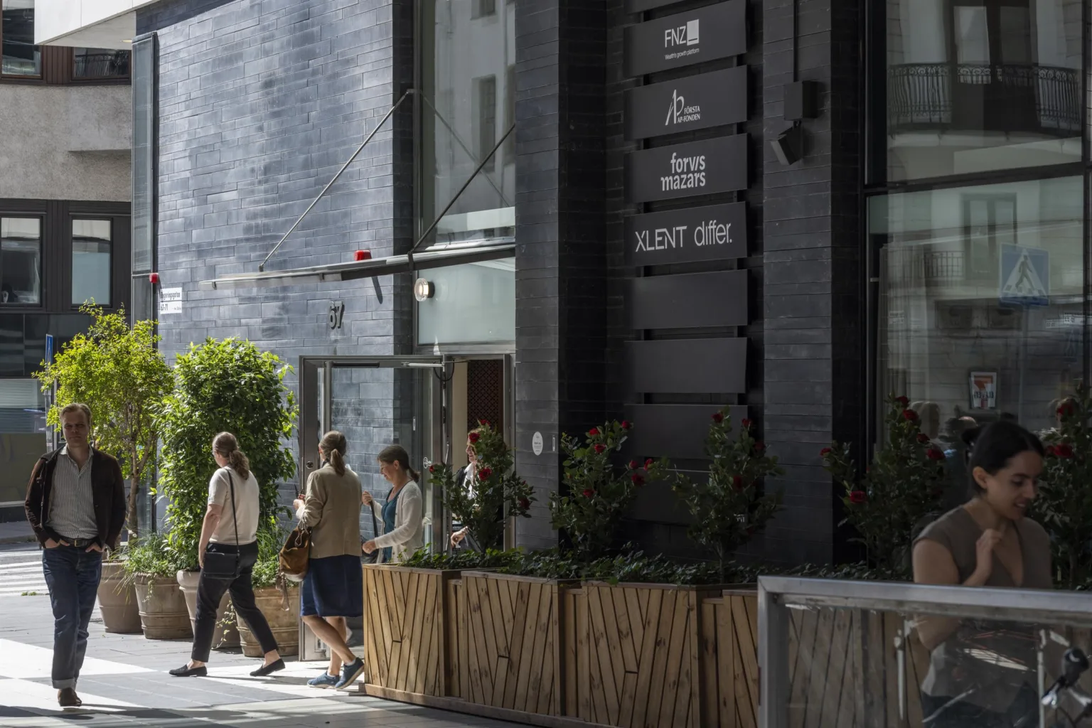 Entré till en byggnad med glasdörrar, växter i träkrukor och personer som går förbi.