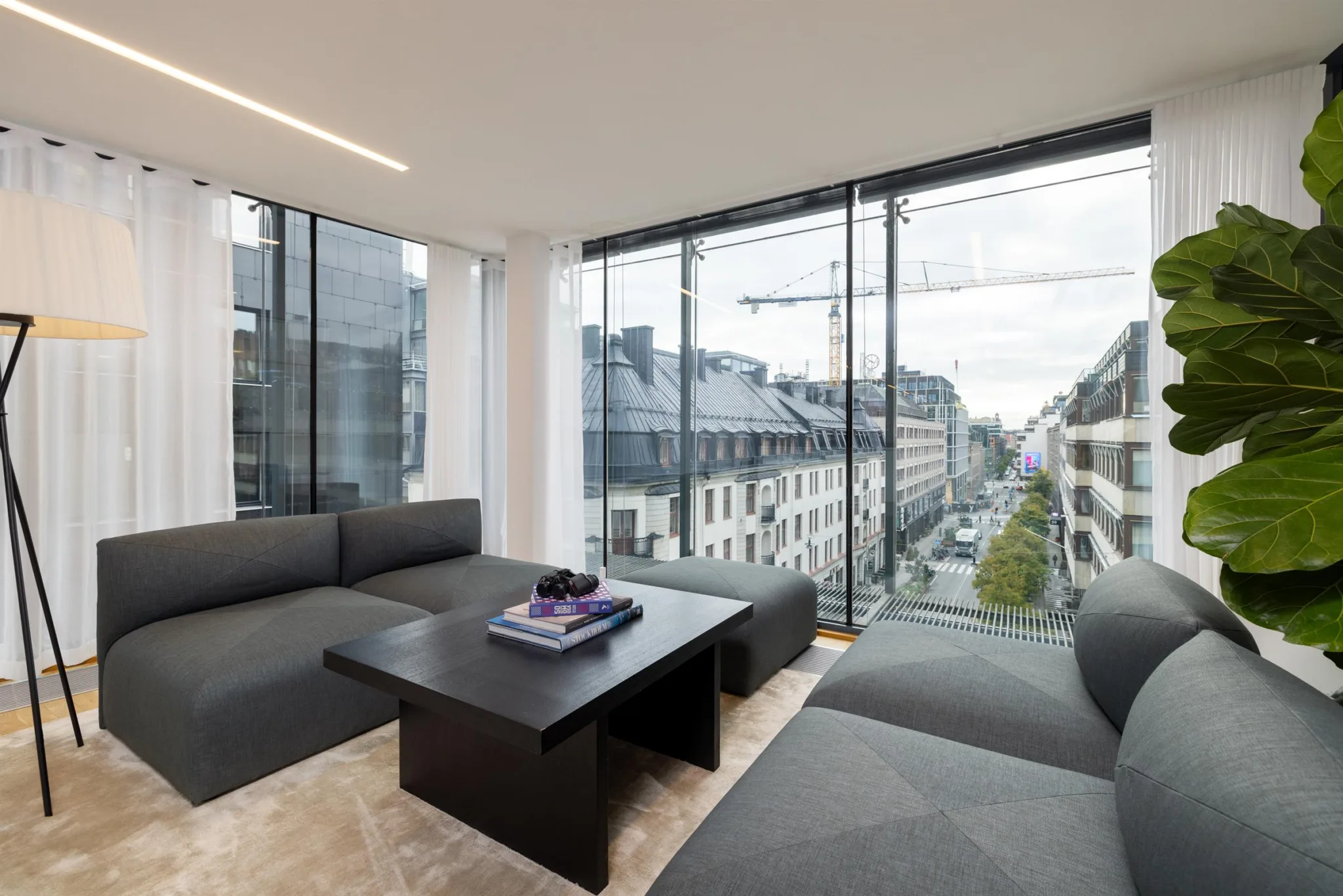 3. En lounge med gråa soffor, ett svart bord och stora glasfönster med utsikt över stadens byggnader.