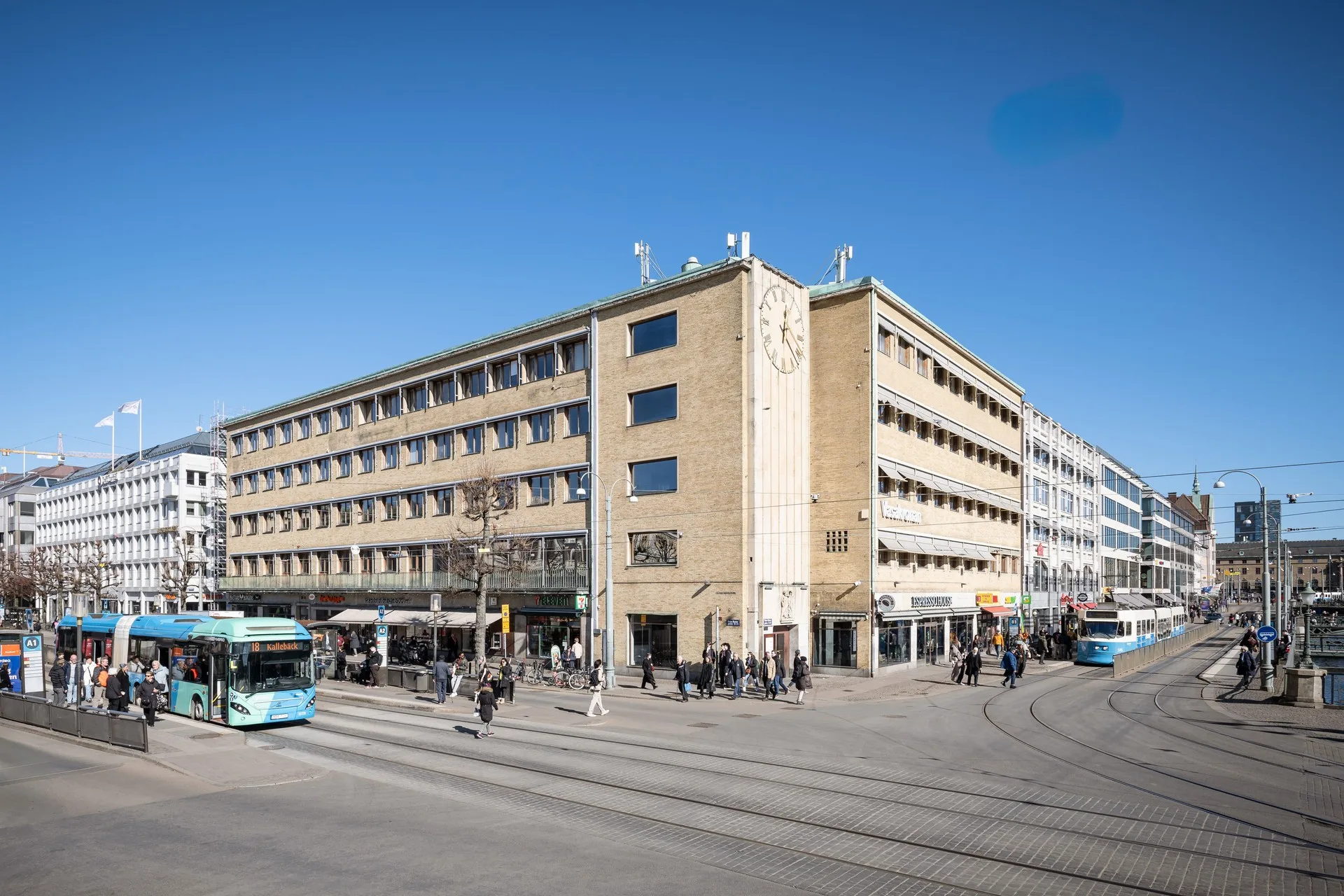 En stor byggnad i tegel med flera våningar, omgiven av människor och spårvagnar på en öppen gata under en klarblå himmel.